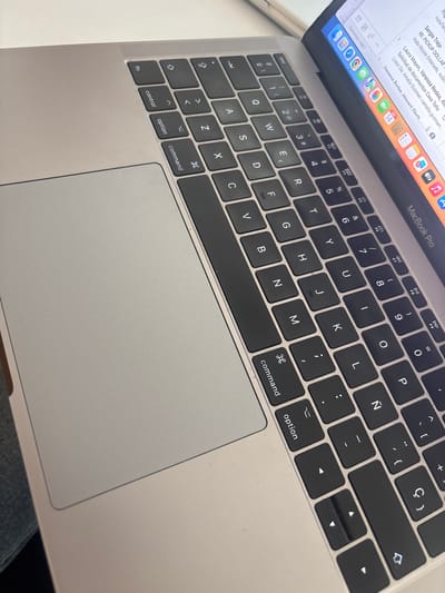 This image is a close-up of a silver MacBook Pro keyboard and part of the screen. The laptop is open, and the screen shows a partially visible email inbox with several email subject lines. The keyboard layout is visible, featuring a Spanish language configuration including the Ñ key. Keys such as "command," "option," "control," "fn," and the arrow keys are clearly shown. The trackpad is also visible. The laptop appears to be resting on someone's lap, and blue jeans are visible along the bottom edge.