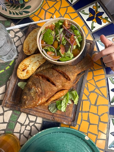 A fried whole fish on a wooden board accompanied by a green salad with tomatoes and red onions, and two grilled eggplant slices. A hand is pointing at the fish. The arrangement is placed on a colorful mosaic tile table. Other items visible include a glass of water, plates, and a glass of amber liquid. The composition appears to be an overhead shot of a restaurant meal.