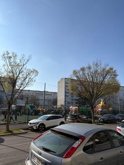 The image captures an outdoor urban scene in Bron, France, under a clear blue sky on a sunny day. In the immediate foreground, the rear of a silver Ford Focus hatchback (license plate "AP-640-HY") is visible on an asphalt street. Several other vehicles are parked parallel to the street, including a white modern crossover, a dark brown compact SUV, and a black hatchback.
Behind the parked cars, a black metal fence encloses a green artificial turf area. Within this enclosure, multiple large, brightly colored inflatable structures are erected, indicating a recreational event or temporary playground. Prominent among these are a multi-level green, yellow, and red inflatable structure on the left, and an orange, yellow, and brown castle-like inflatable structure with a white figure on top, situated on the right. Numerous individuals, appearing to be both adults and children, are present within this fenced area, moving around and interacting with the inflatable attractions. A tall net, consistent with a volleyball net, is stretched across a section of the enclosed area.
Two deciduous trees with nascent green foliage, indicative of springtime, are positioned on either side of the view into the fenced area. The background is dominated by several large, multi-story residential apartment buildings constructed of light-colored materials, featuring multiple windows and balconies. One taller building stands prominently on the right side of the background.