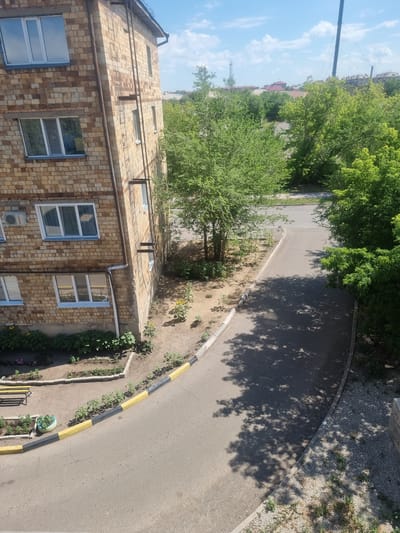 The image depicts an outdoor scene in the city of Saran, Kazakhstan, on a clear day with scattered clouds. In the foreground, a section of an apartment building constructed with light brown brick is visible, featuring several windows with white frames. Adjacent to the building is a paved pathway or road with a yellow and black striped border. The pathway curves away into the distance, where it is partially covered by the dappled shadows of trees. Green trees and some smaller plants are scattered throughout the scene, with a small garden area containing sunflowers and other greenery located near the base of the building. The overall environment suggests a residential area within the city.