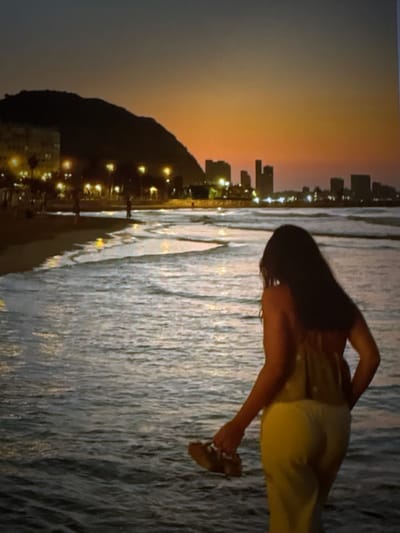 The image shows a person in a yoga pose on a beach at sunset. The person is silhouetted against the colorful sky and the dark water. The beach appears to be rocky, with waves crashing on the shore. In the background, there are city lights and buildings. The overall mood of the image is serene and peaceful. The time of day is sunset, as indicated by the warm colors in the sky. The weather appears to be calm, with no strong winds. The location is a beach, likely in a city, given the presence of buildings in the background. The person is dressed in yoga attire and appears to be practicing a difficult pose. The image is taken from a low angle, looking up at the person and the sky.