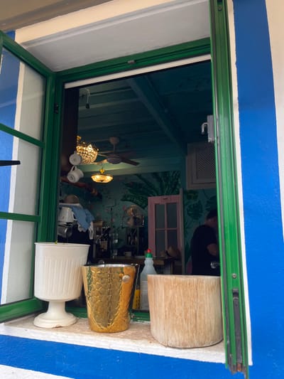 A window with a green frame is open to a view of a cafe interior. The interior is painted a light blue with a wood ceiling. Three light fixtures hang from the ceiling, with a ceiling fan in the center. The walls are decorated with green painted foliage designs. In the foreground, a white ceramic pot sits on the window sill, next to a gold metal ice bucket. A wooden chopping block rests on the windowsill, next to a half-empty plastic bottle. A man, wearing a dark blue shirt, is visible through the window, but his face is obscured. There are several other indistinct objects in the interior view. The cafe is located in Lomo Quiebre, Spain.