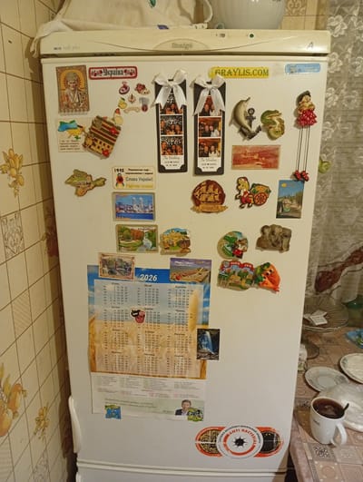 The image depicts a refrigerator door covered in various magnets and a calendar. Several of the magnets feature images of Ukrainian landmarks and symbols, including the national flag, St. Nicholas, and cityscapes. There are also novelty magnets, such as a dolphin, a frog, an elephant, and a character with dangling legs. A calendar for the year 2026 is prominently displayed, showing the days and months in Ukrainian. A small framed photograph of what appears to be a wedding celebration is also visible. On the lower right of the refrigerator, there are two circular stickers; one is a bright yellow with a black asterisk and text that appears to be a slogan related to antibacterial protection, and the other is a circular sticker with a black border and an abstract design. To the right of the refrigerator, a table is set with plates, a cup of tea, and a patterned tablecloth. The overall scene is domestic and suggests a personalized space with collected mementos and practical items. The question of its location in Myrhorod, Ukraine, cannot be answered from the visual information provided.