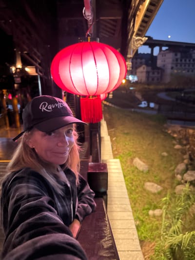 The photograph depicts a person wearing a dark-colored baseball cap with white cursive writing ("Rawmez") and a plaid shirt, leaning against a wooden rail or bench. A red Chinese lantern hangs nearby, illuminated and casting a soft glow. The background shows a street in Zhuoshui, China, with buildings, vegetation, and outdoor lighting. The sky is visible with a partial moon. The photograph appears to be taken at dusk or night.
