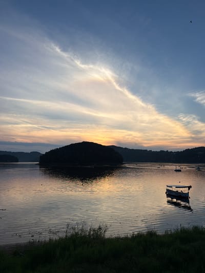 The image depicts a serene sunset over a body of water, likely a lake or a wide river, at Gródek nad Dunajcem, Poland. A silhouetted, tree-covered island is prominent in the middle ground, casting a reflection on the calm water. The sky is a canvas of soft blues, oranges, and yellows, with wispy clouds stretched across it, indicating the sun is either setting or has just set. In the foreground, the dark silhouette of grass and reeds lines the shore. Several boats are scattered across the water, some are small, while one larger boat with a canopy is anchored in the right mid-ground, its reflection also visible. The overall atmosphere is tranquil and picturesque.