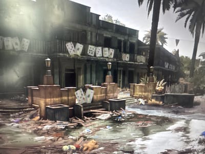 The image depicts a grimy, dilapidated urban scene, likely set in a tropical or subtropical climate due to the presence of palm trees. The central focus is a two-story building with balconies adorned with large playing cards, including what appear to be Aces and other suit cards. The lower part of the building has a weathered facade with boarded-up windows. In front of the building, the ground is littered with debris, including trash, broken wood, and puddles of water, suggesting recent flooding or poor sanitation. Several large, square, rustic containers and planters are scattered around the foreground. A streetlamp stands in front of the building. The overall atmosphere is one of decay and neglect. There is no visible indication or context suggesting this scene is in the city of Stepnogorsk, Kazakhstan.