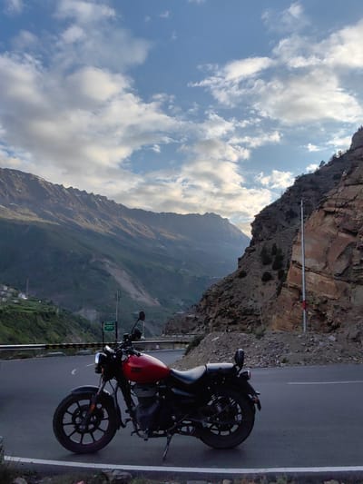 A red Royal Enfield motorcycle is parked on the side of a paved mountain road in Stingri, India. The motorcycle is positioned in the foreground, facing left, with its front wheel turned slightly. Behind the motorcycle, the road curves to the left, bordered by a metal guardrail. The setting is a dramatic mountain landscape with steep, rocky cliffs on the right and a vast, verdant valley stretching into the distance on the left. The valley is dotted with small villages and terraced fields, and the distant mountains are shrouded in a hazy, soft light.
The sky is a vibrant blue with scattered white clouds, suggesting it is late afternoon or early evening. The lighting casts a warm glow on the landscape, highlighting the textures of the rocks and the lush green slopes. The overall mood is one of serene adventure and the beauty of nature. A green signpost is visible near the motorcycle, though the text is not entirely legible in the original image. A tall metal pole with red and white markings stands near the rocky cliff. The scene evokes a sense of freedom and exploration, typical of motorcycle touring in mountainous regions.