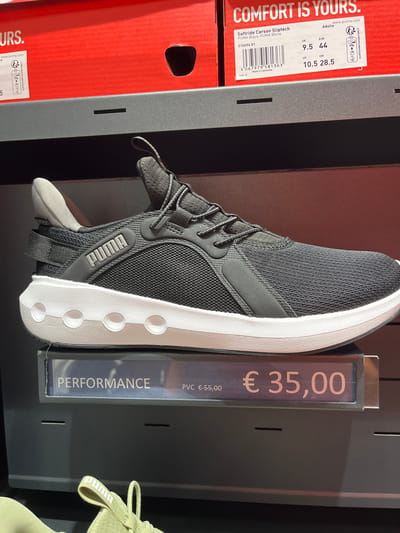 The image depicts a retail display of Puma shoes in what appears to be a store in Rodengo, Italy. In the foreground, a black Puma softstride sneaker with a white sole is prominently displayed on a shelf. To the left of the shoe, a price tag reads "PERFORMANCE € 55,00" and to the right, a larger price tag displays "€ 35,00". Above the shoe, a portion of a red Puma shoe box is visible with text that includes "COMFORT IS YOURS." and a product label for "Softride Carson Sliptech PUMA Black-PUMA White" with size information. The environment is an indoor retail setting, likely a shoe store or a department store with a shoe section. There are no people visible in the image.