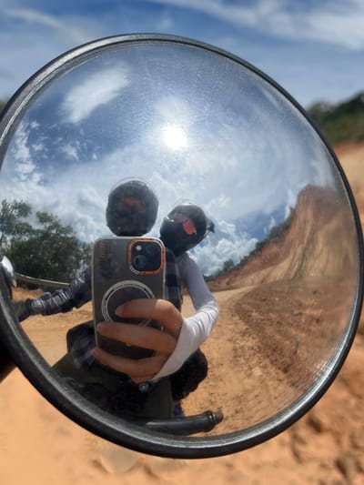 The media file is a reflection in a round convex mirror, likely a motorcycle mirror, capturing a selfie. The main subject is a person wearing a black helmet with orange and red accents visible through the visor, and a grey and white long-sleeved shirt. The person is holding a smartphone in their right hand, using it to take the photo. Their left hand is also visible, with a white sleeve.
The reflection shows the person from the chest up, with another helmeted figure, possibly a passenger or another rider, reflected behind them. The background of the reflection is a sunny outdoor scene. The sky is blue with white clouds, and the sun is bright, suggesting it is daytime. The ground appears to be a sandy, dirt track, with some dry vegetation and sparse trees on the right side of the frame. The terrain looks somewhat arid and uneven, consistent with a rural or off-road location. The overall mood appears to be casual and adventurous, given the context of outdoor exploration.
The smartphone in the reflection has a dark case with orange trim around the camera lenses and some circular white graphics. There are faint markings on the case which are not clearly legible. The mirror itself has a black rim and appears to be slightly dusty. The setting suggests a location in Vietnam, possibly Son Dien, given the context.
