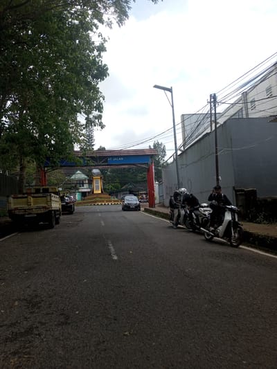 The image shows a road with a "AT JALAN" sign arching over it. There is a monument in the middle of the road, and several vehicles are visible, including a truck and some motorbikes. Three people are standing near the motorbikes, and a few other people can be seen further down the road. The environment includes trees, a building, a wall, and overcast skies. It appears to be a road in a city. It is not certain if the location is Ban Phouviengnoy, Laos.