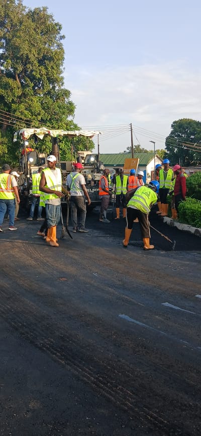 A group of construction workers are repairing a road in Jama A, Nigeria. The workers are wearing high-visibility vests and safety helmets. They are using a paving machine to lay down asphalt. The scene appears to be a work in progress, with fresh asphalt being spread and smoothed. The sky is clear and the sun is shining, suggesting it is daytime. Trees and buildings can be seen in the background, indicating an urban or semi-urban environment. The overall impression is one of ongoing infrastructure development.