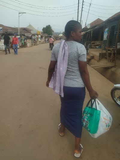 The image shows a woman walking down a paved street, carrying a lavender scarf and a green bag, with a white plastic bag in her other hand,  in the city of Junction, Nigeria. The woman is wearing a grey shirt, a blue skirt, and sandals. The street is lined with buildings, and there are some power lines overhead. In the background, another woman is walking in the opposite direction, and a man is riding a motorcycle. The scene is bright and sunny.