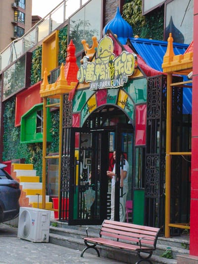 The image shows the exterior of a brightly colored playground or entertainment venue in Jiguan District, China. The facade features a whimsical design with a cartoon duck mascot, a blue domed roof, and walls adorned with red, yellow, and green stained-glass-like panels. The entrance is framed by black metalwork and a dark doorway. To the left of the entrance, a set of yellow and white stairs leads up to a green structure. An outdoor air conditioning unit is visible on the ground near the stairs. A dark-colored car is parked to the left of the stairs. In front of the entrance, a pink bench sits on a grey concrete walkway. Two individuals are partially visible through the doorway, with one appearing to hold something. The overall impression is one of a vibrant, family-oriented attraction.