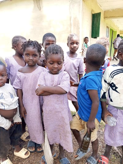 A group of young African children are standing in front of a weathered building with a green metal window in Andaha, Nigeria. The building appears to be a school. Many of the children are wearing matching purple and white checkered dresses. One child in the foreground is wearing a blue polo shirt and khaki shorts, holding a dark bottle. Another child is holding a clear plastic water bottle. The ground is covered with dirt and scattered debris. The overall atmosphere suggests a casual outdoor gathering or school activity.
