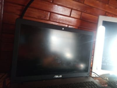 The image shows a close-up view of a laptop screen and a portion of another screen, likely a tablet or monitor, to the right. The laptop is black with the "ASUS" logo visible on its lower bezel. The screen of the laptop is dark and reflective, showing some smudges or dust. Above the laptop screen, a small, square camera lens is visible. The background consists of a wall paneled with wooden planks, which are polished and have a warm, reddish-brown hue.
To the right of the laptop, another screen displays a bright interface with a clock showing "9:23" and text that appears to be in Cyrillic, possibly indicating a date and time. The right screen is illuminated by a bright light source, causing a glare. Cables are visible in the foreground, suggesting an electronic setup. The overall scene is indoors, with the wooden wall suggesting a rustic or cozy environment. There are no people visible in the image. The time appears to be either morning or evening, based on the "9:23" on the secondary screen. The lighting and the reflective surfaces suggest the image was taken in an environment with artificial lighting, possibly in Piskivka, Ukraine, as per the location context.