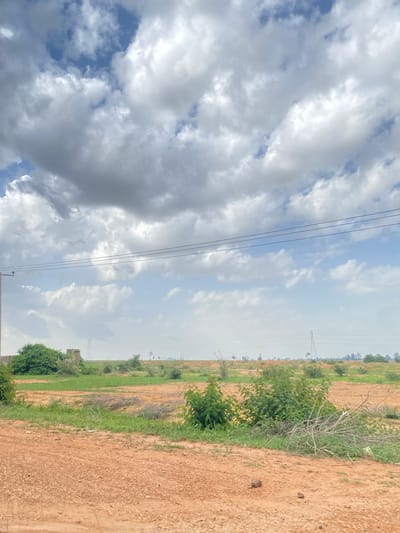 This image captures an outdoor scene in Barakin Kuru III, Nigeria, under a cloudy sky. The frame is divided diagonally. On the left side, a dramatic sky filled with heavy, dark gray clouds and patches of blue is visible, with thin black electrical wires stretching horizontally across it. The right side of the image depicts a rural landscape. A dirt road or path runs along the right edge, its surface composed of reddish-brown soil with small stones. Adjacent to the road, a strip of green grass and low-lying vegetation marks the transition to a field. In the distance, scattered trees and shrubs dot the landscape, with a tall, lattice-like electrical pylon visible further back. The overall impression is of an overcast day in a rural, possibly developing, area. There is no visible text in the image.