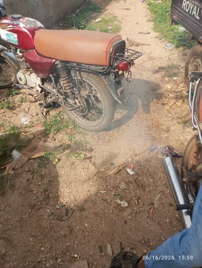 In Dokan Tofa, Nigeria, a rusty red motorcycle is parked on a dirt path. The motorcycle has a brown seat and a visible exhaust pipe that is emitting smoke. To the right of the motorcycle, a portion of a dark-colored truck with the word "ROYAL" visible on its side is present. The ground is covered with dirt, small stones, dry leaves, and scattered pieces of white plastic. In the foreground, a person's blue jeans and a dark shoe are visible, along with a date and time stamp of "06/16/2026 15:59".