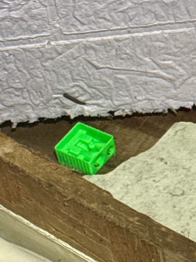 This is a close-up, overhead shot of a small, bright green, cube-shaped object with a lattice-like pattern on its surface. It appears to be a Lego brick or a similar interlocking building toy. The object is positioned on a textured, light-colored surface, possibly a wall or a floor with a plaster-like finish. To the left of the green object, there is a torn piece of a white, crumpled material that has a wrinkled, almost fabric-like texture. A thin, dark, elongated object, possibly a twig or a dark fiber, lies across this white material. To the right of the green object, and behind it, is a dark, shadowy area that transitions into a rough, brown, wooden surface. This wooden surface appears to be part of a larger object or structure, possibly a beam or a piece of furniture, with visible grain and texture. The lighting suggests an indoor environment, with a soft, diffused light source illuminating the scene. There is no discernible text in the image. The scene appears to be an everyday setting, possibly a home or a workshop, with no specific activity or event taking place beyond the placement of the object. The focus is on the textures and colors of the materials, with the bright green object serving as a focal point against the more subdued background.