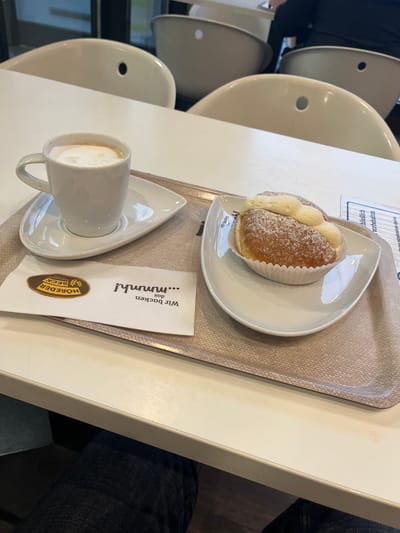 The image, rotated 90 degrees counter-clockwise for natural viewing, captures a close-up of a cafe or bakery table setting. On a white table, a beige textured tray holds a small white ceramic cup of foamy coffee, likely a cappuccino or latte macchiato, placed on a matching leaf-shaped saucer.
Next to the coffee, a white paper napkin is visible, featuring the German text "Wir backen das ...mmmh!" and the brand "HÖRDER BECK" accompanied by a logo resembling an ear of wheat. Further along the tray, a similar leaf-shaped white plate holds a pastry. This pastry, resembling a Krapfen or Berliner, is golden-brown, dusted with powdered sugar, and appears to be filled with cream, resting in a white pleated paper liner.
In the background, several minimalist white chairs are partially visible and out of focus, suggesting an indoor public seating area. The lower right corner of the image shows a person's leg clad in blue jeans, seated at the table. Faint, inverted German text, including "Alle Rechte vorbehalten" (All rights reserved), can be seen on a paper or sign in the upper left background. The visible German text and the specific type of pastry suggest the scene is set in a German bakery or cafe, consistent with the location context of Bamberg, Germany. The overall impression is one of a relaxed coffee and pastry break.