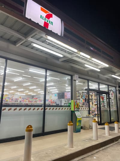 The image depicts a 7-Eleven store exterior in Baan Kai, Thailand. The store has a brightly lit interior visible through large glass windows. The 7-Eleven logo is prominently displayed on the exterior. A row of white and gold bollards are located in front of the store, and green and blue waste bins are seen on the pavement. A "WIFI" sign is visible on the exterior wall. The interior shelves are stocked with various products, and signage for "All Cafe" and "Ready To Eat" is visible.