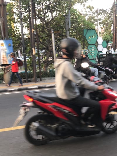 The image shows a street scene in Sidoarjo, East Java, with several people on motorcycles. In the foreground, a person wearing a gray long-sleeved shirt and a black helmet is riding a red motorcycle. Behind them, another person on a motorcycle, wearing a black jacket and a helmet, is visible. Further in the background, more people on motorcycles can be seen. The setting appears to be a roadside with trees and some greenery. There are also some hexagonal-shaped structures visible in the background, possibly part of a building or advertisement. The lighting suggests it is daytime. The overall impression is a busy street with motorcycle traffic. There is no visible text in the image.