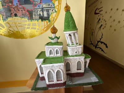 The image depicts a miniature model of a church with green onion domes, situated on a table. The model is constructed with white and red elements, resembling bricks and a dark red base. The roofs are green and textured, mimicking shingles. The church has multiple levels with arched windows. In the background, a wall is visible with a yellow-beige paint. A large circular mural is present on the wall, featuring gold foil and images of buildings and a statue. To the right of the mural, another section of the wall displays a brown-colored illustration of dandelions scattering their seeds. Text in Russian is also visible on the wall, partially obscured by the mural. The scene appears to be indoors, likely in a public space or educational institution, given the decorations on the wall. There are no people visible in the image.