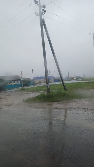 The image depicts a rural street scene in the village of Kiyasovo, Russia, under overcast and misty conditions. A weathered wooden utility pole with multiple crossbars and insulators stands prominently in the foreground, leaning slightly. Two parallel support poles are visible. The road in the foreground is wet and reflective, indicating recent rain, with puddles and a dirt shoulder. Beyond the road, to the left, is a traditional wooden house with a blue fence. Further down the street, other rural houses and structures are visible, some with blue roofs. A yield sign and a pedestrian crossing sign are present near the road. The overall atmosphere is somber and damp, characteristic of a foggy or rainy day in a village setting.