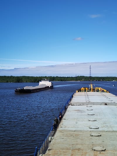 The image depicts a waterway, likely a river, with two barges visible under a partly cloudy blue sky. The barge closer to the viewer appears to be a flat-deck barge with a gray surface and blue railings. A second barge is positioned further away, consisting of a dark hull carrying a covered load and a white superstructure. Trees line the horizon, and a tall metal transmission tower can be seen in the distance. The location is identified as Novodevich'e, Russia. There are what appears to be a few figures on the deck of the flat-deck barge closer to the camera.