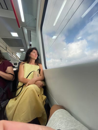 A woman with dark hair, wearing a yellow dress, sits on a train, looking out the window at the clouds. She is holding a woven bag. The train interior is visible, with seats, overhead lights, and a red pole. Another passenger, wearing a maroon shirt, is visible in the foreground. The train is likely traveling through or near Sant Andreu de Llavaneres, Spain, given the context provided. The view outside the window shows a bright sky with scattered clouds.