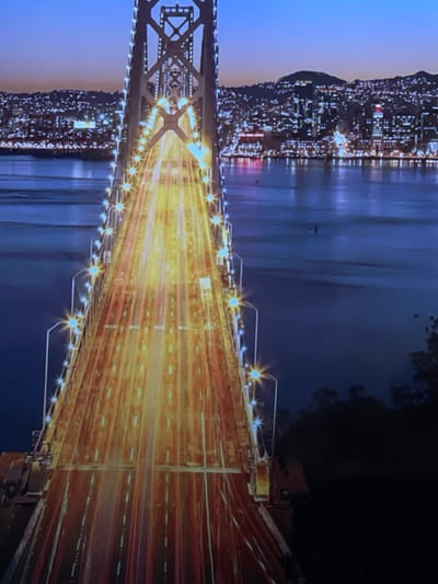 The image depicts a nighttime scene in Nizhnyaya Urat'ma, Russia, featuring a large, illuminated suspension bridge. The bridge's multi-lane roadway extends from the lower foreground towards the upper center, showcasing elongated streaks of yellow and red light, indicative of vehicle movement captured with a long exposure. Numerical "80" markings, likely speed limits, are visible on the road surface. The bridge is lined on both sides by numerous streetlights, appearing as bright, star-like sources. A prominent metal lattice tower, part of the bridge's support structure, rises towards the top center, also adorned with lights. The bridge spans a wide, dark body of water. On the far side of the water, a dense urban landscape is visible, characterized by a multitude of scattered artificial lights extending up a hillside against a dark twilight sky. The lower right portion of the image shows dark, indistinct foliage near the bridge's base. The perspective is elevated, looking down the length of the bridge towards the distant cityscape.