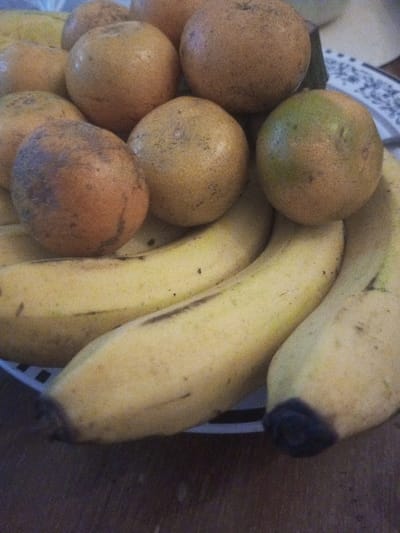 The image displays a close-up, slightly overhead view of ripe fruit arranged on a white plate with a dark, patterned edge, located in Karaikudi, India. The composition primarily features numerous round citrus fruits, identifiable as oranges or mandarins, piled atop a curved arrangement of yellow bananas. The citrus fruits display a spectrum of yellowish-orange and light brown colors, with visible dark blemishes and small spots; one fruit in the mid-right foreground possesses a noticeable green discoloration. The bananas are ripe yellow, marked with small black spots and dark streaks, and at least two visible banana ends are discolored to a dark brown or black. The plate rests on a dark brown, textured surface, consistent with wood grain. No individuals, actions, or interactions are observable within the frame.
