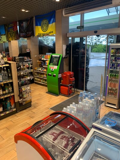 The image shows the interior of a convenience store in Solonytsivka, Ukraine (latitude: 49.973, longitude: 36.02). The store is relatively small, with wooden floors, and a variety of goods on display.  Two glass-front refrigerators sit in the foreground, one containing packages of frozen food, the other empty. A large green ATM machine is visible in the center of the image. To the left, a wall of shelves displays a variety of alcoholic beverages, including bottles of vodka, wine, and beer. A Ukrainian flag hangs on the wall behind the ATM machine.  A glass door marked "вихід," meaning "exit" in Ukrainian, is visible in the background, allowing a view outside the store. The entrance to the store is just beyond the ATM machine, and the door to the right of the ATM machine is open.  A person can be seen standing behind the counter. The store appears to be well-lit, and the temperature appears to be moderate.