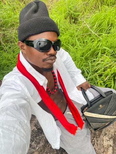 An overhead, diagonally oriented shot captures a person lying outdoors, looking up towards the camera. The subject, appearing male, has dark skin, a short beard, and is wearing black sunglasses and a black knitted beanie. They are dressed in a white long-sleeved garment, possibly a tunic or shirt, over gray trousers. A bright red, fringed scarf or fabric piece is draped around their neck, partially revealing a black and red beaded necklace. They hold a black handbag with a light-colored stripe and metallic accents in their right hand. The background features vibrant green grass, indicating a natural outdoor setting. The person appears to be resting on a patch of uneven, brownish-gray ground, possibly sand or rock. The bright lighting suggests it is daytime. The scene is located in Afikpo, Nigeria.