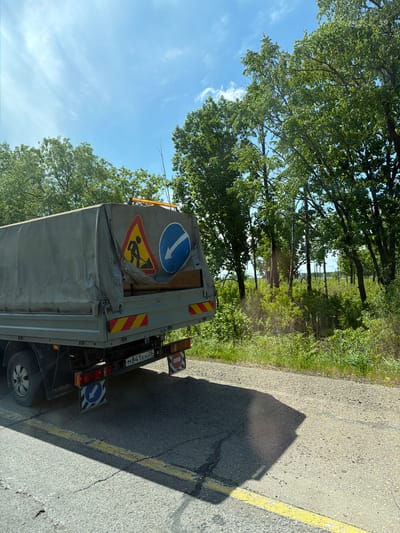 A utility truck with a gray tarp covering its cargo bed is parked on the right side of a cracked asphalt road in Aursky District, Russia. The truck displays several warning signs, including a yellow triangle with a black silhouette of a person digging, and a blue circular sign with a white arrow pointing left. A yellow and red striped reflective marker is visible on the rear bumper, along with a license plate that reads "M841KH 28". The truck is casting a long shadow on the road. The background features dense green trees and foliage under a bright blue sky with scattered clouds. The environment appears to be a rural or semi-rural area.