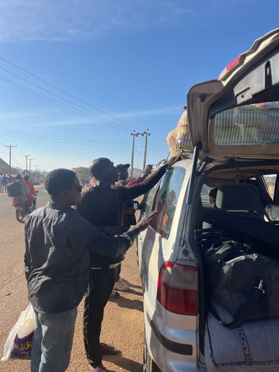 Two people secure luggage atop a car on a sunny day. One person wears a grey jacket and jeans, while the other is in a black top and pants. The car trunk is open, revealing more luggage inside. Power lines stretch across the clear blue sky. Other people and motorcycles are visible in the background on what appears to be a road.