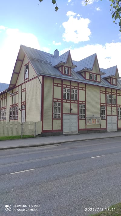 The image shows a two-story wooden building with a metal roof, located on a street in Iisalmi, Finland. The building has a light yellow facade with dark red trim around the windows and along the architectural details. It features multiple windows on both floors, some of which are grouped together. There are several dormer windows on the roof. In front of the building, there is a light-colored wooden fence. A road is visible in the foreground, with markings on the asphalt. There are some posters displayed on the building's facade. The sky is blue with some white clouds, and tree leaves are visible in the upper right corner of the image. The building's signage reads "TYÖVÄENTALO".