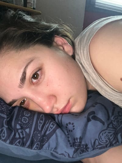 This image captures a young adult female in a reclining position, indoors, located in Ancenis, France. Her head is positioned on a blue pillow displaying a dark paisley pattern. She lies on her right side, with her face oriented towards the camera, her chin resting on the pillow's surface. Her brown eyes are open and maintain a direct gaze. Dark hair is visible along the top of her head. She is clad in a light-colored tank top, which reveals her left shoulder and a portion of her upper chest. A small, dark mole is discernible on her upper chest. The background is out of focus, with a shelf holding containers visible on the upper left, and a window with horizontal blinds visible on the upper right.
