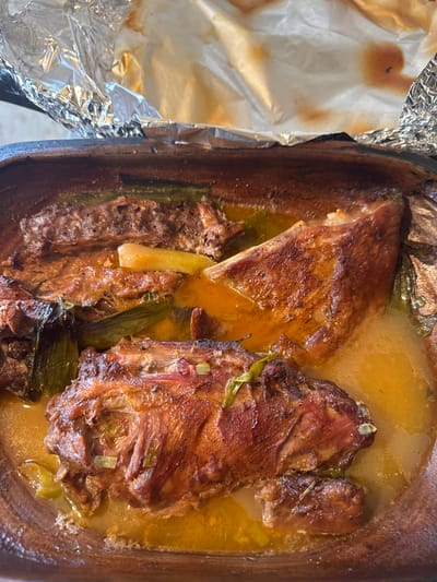 The image shows cooked meat, possibly pork or lamb, in a large, shallow, brown clay pot. The meat appears to be browned on the outside and is immersed in a yellow-orange broth. There are also green, stalk-like vegetables (possibly celery or leeks) scattered around the meat. A piece of aluminum foil is present on the left side of the pot. The image appears to be taken from an overhead perspective. There is no definitive information in the image itself to confirm its location in Novi Chiflik, Bulgaria.