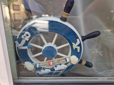 The image shows a decorative ship's wheel with a nautical theme, positioned on a window. The wheel is predominantly white and blue, with rope accents and attached decorative elements like an anchor, shells, fish, and a seagull. The wheel is situated behind a window pane, with a light-colored screen or mesh overlaying the view through the glass. Reflections from an outdoor environment, possibly buildings and trees, are visible on the window's surface. The overall setting suggests an interior space looking outwards through a window. This scene is located in Antony, France.