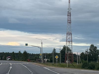 The image depicts a scene on a road in the village of Babanaovo, Russia, under a cloudy sky. A tall, red and white lattice tower, likely a telecommunication mast, dominates the right side of the frame. In the foreground, a paved road curves to the left, marked with white lines indicating lanes and a dashed line for passing. Speed bumps are visible across the road. To the left of the road, several road signs are present, including a pedestrian crossing sign within a blue square and two yellow diamond-shaped signs. A lamppost is also visible. In the background, a dense line of trees forms the horizon, with a few houses with red roofs nestled amongst them. Power lines and utility poles are scattered throughout the landscape. The overall impression is a rural or semi-rural setting on an overcast day.