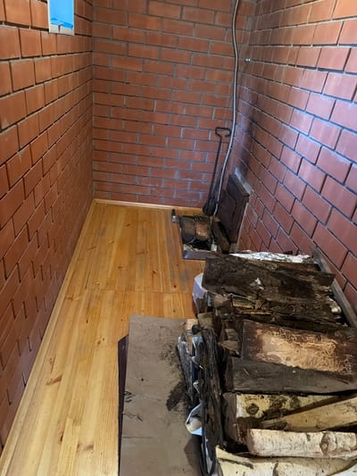 This image shows an interior view of a room with brick walls and a wooden floor. In the foreground, there is a pile of firewood, consisting of several logs. Behind the firewood, there is a metal fireplace tool leaning against the wall. The room appears to be a storage area or a room designed for a fireplace. The lighting suggests it is daytime, possibly with natural light coming from an unseen window or artificial lighting. The overall scene is rustic and practical, consistent with a residential or utility space. The specific location in the city of Каменка, Russia cannot be determined from the visual information alone.