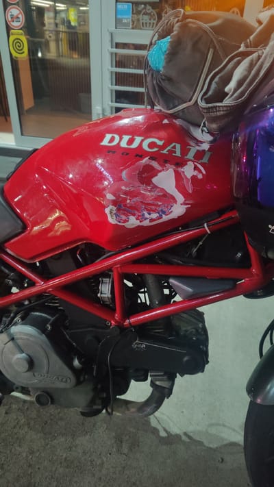 The image shows a red Ducati motorcycle parked outdoors, possibly in a city like Dagomys, Russia, though the location is not explicitly confirmed. The motorcycle is the primary subject, with its red fuel tank prominently displaying the "DUCATI" logo and some scuffs or damage. A brown bag, possibly containing a blue object, is resting on the fuel tank. The motorcycle has a red frame and visible engine components. In the background, there are reflections in glass windows, possibly from a store or building, with signage including "DELTA" and "24/7". The ground appears to be paved. No people are visible in the image.