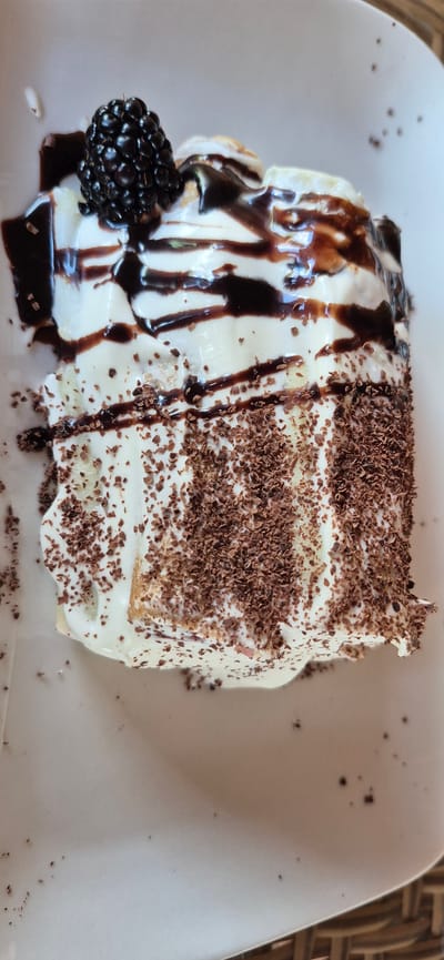A slice of dessert, likely a cake or pastry, is presented on a white plate. The dessert is topped with a generous amount of whipped cream, drizzled with chocolate sauce, and garnished with a single blackberry on top. The sides of the dessert are coated with fine chocolate shavings. The plate is set on a surface that appears to be woven, possibly a placemat or part of a table. The background is a plain, light color. There are no people or any other objects visible that would indicate a specific location in Molovata Nouă, Moldova.