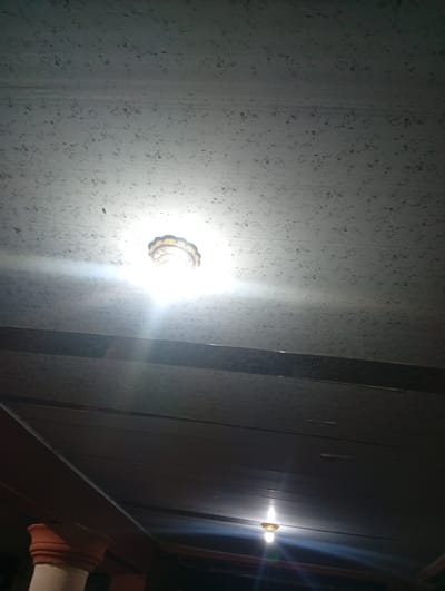 The image shows an indoor scene, likely a room with a textured ceiling. A light fixture is visible on the ceiling, emitting a bright glow that illuminates the surrounding area. There's another light fixture lower down, also on, creating a halo effect. The ceiling appears to be made of panels and has a speckled pattern. In the lower part of the image, a pillar is partially visible, suggesting an architectural element. The lighting is dim, with the light fixtures being the primary sources of illumination, indicating it could be nighttime or a dimly lit interior. The overall impression is a simple, functional interior space. There is no visible text in the image.