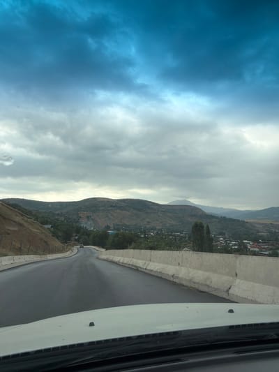 The image depicts a scenic view from a vehicle moving along a paved road in Khumsan, Uzbekistan. The road curves gently to the left, with a concrete barrier lining its right side. The landscape is characterized by rolling hills covered in dry grass and sparse vegetation. In the distance, a small settlement with red-tiled roofs is nestled amongst trees. The sky above is dramatic, filled with a mix of dark, moody clouds and brighter, blue patches. The overall impression is of a rural road leading through a hilly terrain under an overcast sky.