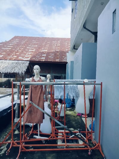 This image, captured in Cottarum Tunong, Indonesia, depicts an outdoor area between two structures, functioning as a storage or work yard. In the immediate foreground and midground, a series of interconnected, orange-painted metal clothing racks on wheels occupy the space. A prominent pale-skinned female mannequin, adorned with a pleated, reddish-brown sleeveless garment, stands on a pole support within the framework of these racks. Behind it, a smaller mannequin head is visible. To the right, a white, headless torso-only dress form is positioned on a stand. A long line of numerous white plastic clothes hangers hangs from one of the metal bars of the rack system. The ground below consists of concrete or pavement, exhibiting patches of green moss or algae, and scattered items including blue plastic bags and white buckets.
In the background to the left, a building with a large, heavily rusted corrugated metal roof is visible, partially obscured by a section of a thatched roof below it. Its walls appear to be constructed from unrendered brick or concrete. To the right, a modern-style, multi-story building with light grey or off-white painted walls and visible windows and a gutter system stands tall, forming a sharp architectural contrast. The scene is illuminated by natural light under a partly cloudy blue sky. No human figures are present, and no active interactions or specific actions are occurring.