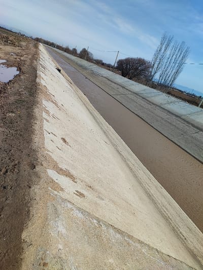 An angled view shows a concrete irrigation canal filled with muddy water. One side has a steep, textured concrete embankment leading to a patch of dirt with puddles. The opposite side is more level. A line of trees and power lines stretches into the distance, with mountains faintly visible on the horizon. The sky is blue with scattered clouds. The lighting is bright and casts shadows along the concrete structure.