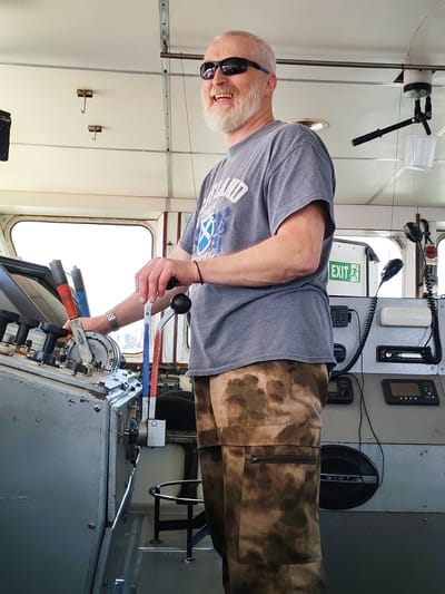 A middle-aged man with a white beard and sunglasses stands at the controls of a vessel. He is wearing a gray t-shirt and camouflage pants. He has his hands on the ship's controls. Behind him are communication devices, a microphone, and an exit sign. The setting appears to be the interior of a ship or boat. There is no evidence in the image to suggest the location is Koprino, Russia.