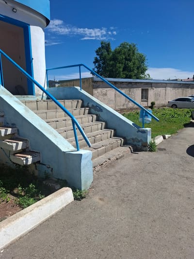 The image shows a concrete staircase with blue railings leading up to a white and blue cylindrical structure. The staircase is in a state of disrepair, with chipped paint and broken concrete. The area around the staircase is covered in overgrown grass and weeds. In the background, there is a long, low concrete building with a barred window and a white car parked in front of it. The sky is blue with a few white clouds. The scene appears to be in an outdoor public space, possibly near an industrial area or a facility. The specific location in the city of Jitiqara, Kazakhstan, is not evident from the image itself, but the overall aesthetic suggests a somewhat utilitarian and potentially older infrastructure.