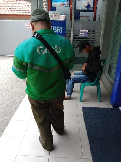 A man in a green "Grab" jacket and brown pants stands, facing away, wearing a crocheted hat. Another man sits on a turquoise chair beside an ATM entrance. He wears a black hoodie, ripped jeans, and a cap. The setting appears to be an outdoor ATM waiting area. A blue doormat lies on the tiled floor next to a blue panel. Advertisements are displayed above the ATMs. A sign in Indonesian language is visible.